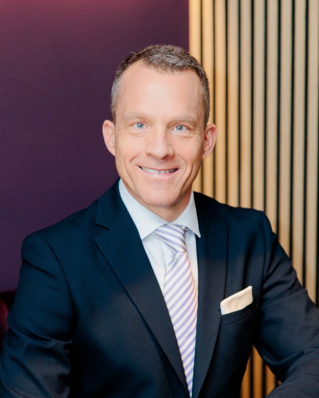 Porträt von Jens Bolms im dunklen Anzug, sitzend vor einer Wand aus Holzlamellen
